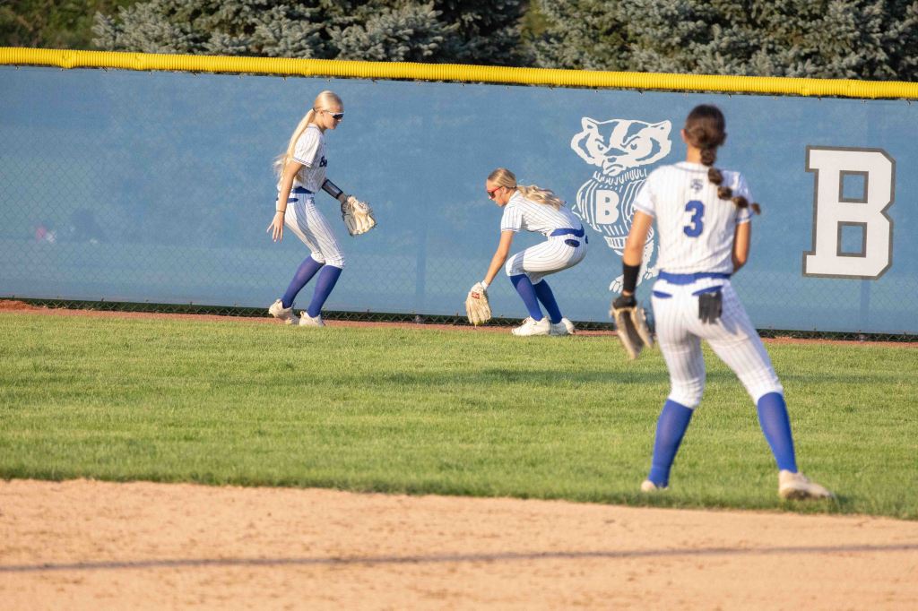 Three female softball players in a field preparing to catch a fly ball, with a blue and white backdrop featuring a badger mascot and a letter B.