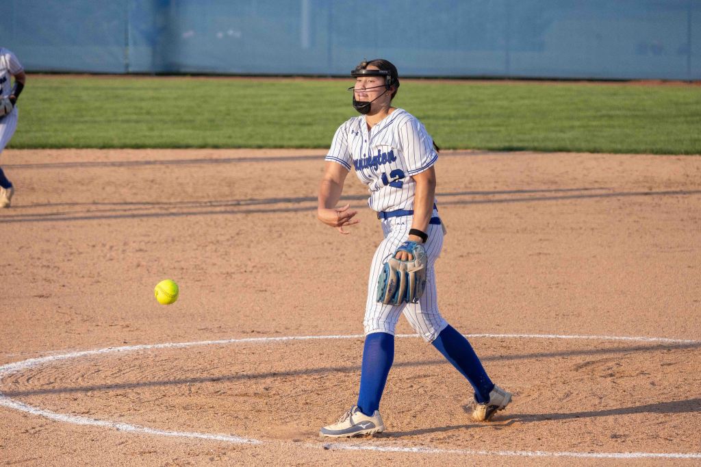 A softball pitcher in a striped uniform releases a yellow softball during a game, with a grassy field in the background.