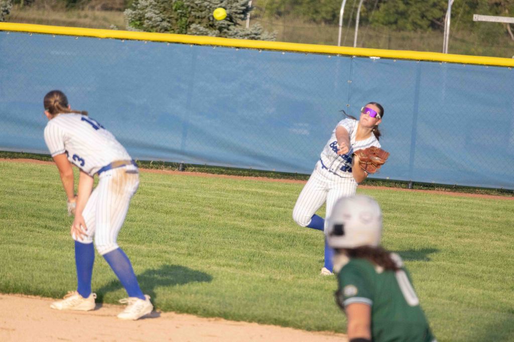 A softball player in a striped uniform throws the ball while another player prepares to catch it. The background shows a blue fence and grass.