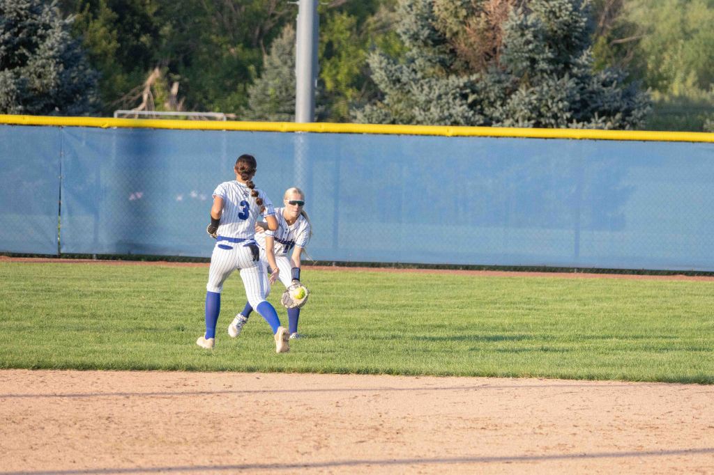 Two female softball players in blue and white uniforms are positioned in an outfield setting. One player is fielding a ball while the other is preparing for a catch, with a baseball diamond and a fenced area in the background.