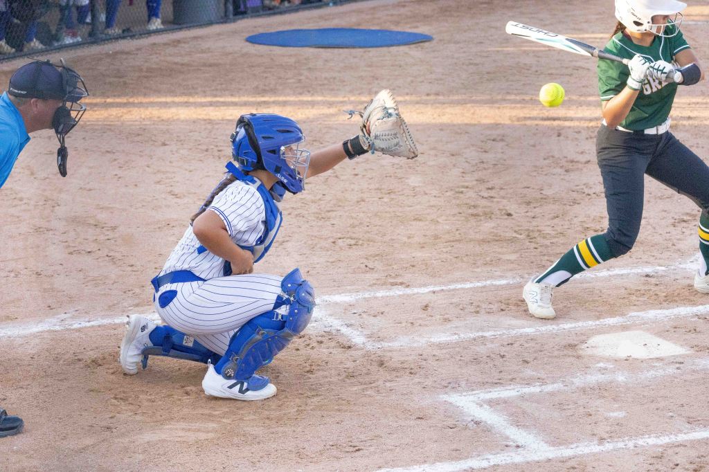 A catcher in blue gear squats behind home plate as a batter in green swings at a pitch during a softball game.
