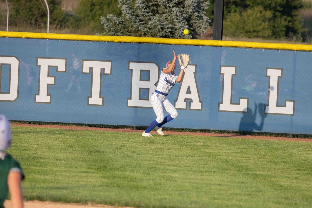 A softball player in a blue and white striped uniform is catching a fly ball near a outfield fence that has 'SOFTBALL' written in large letters.