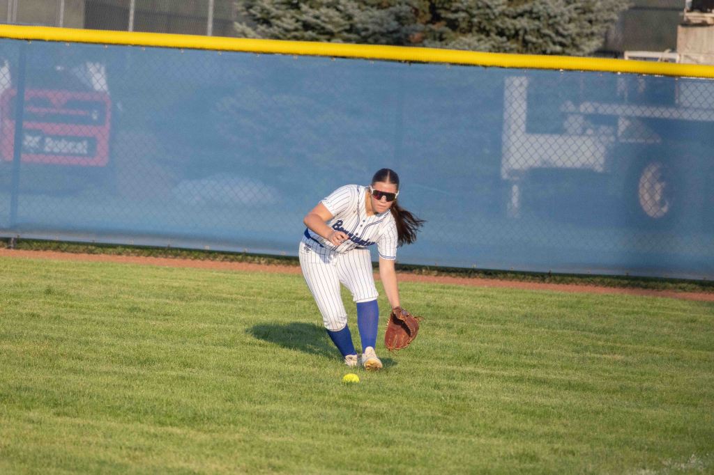 A female softball player in a striped jersey and blue pants crouches to catch a ball on the field.