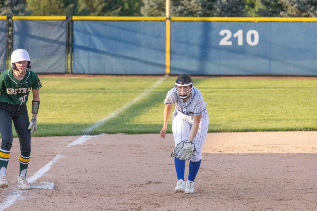 A female softball player in a white and blue uniform crouches in a ready position on the field, while another player in a green uniform stands on base nearby.