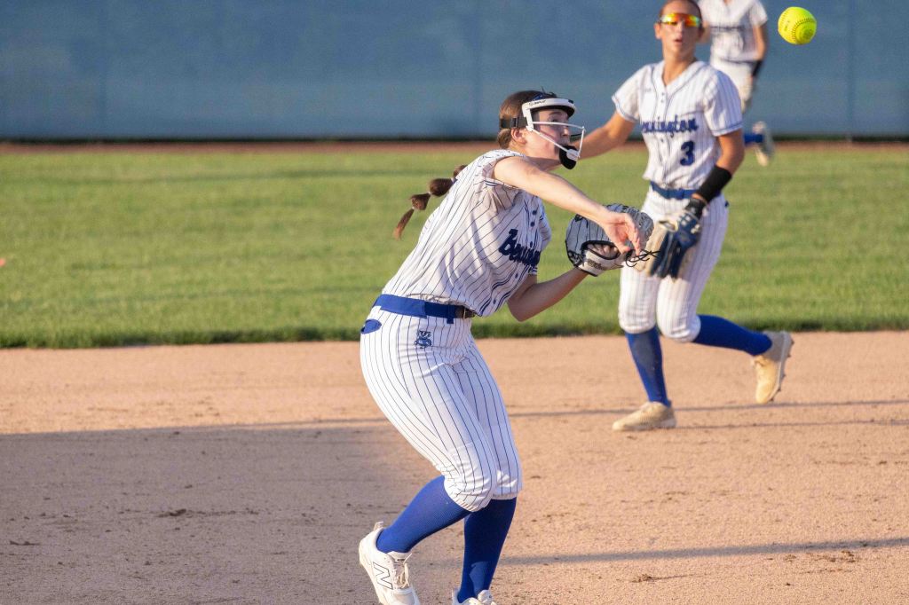 A young female softball player in a striped uniform throws a ball during a game, with another player in the background watching.