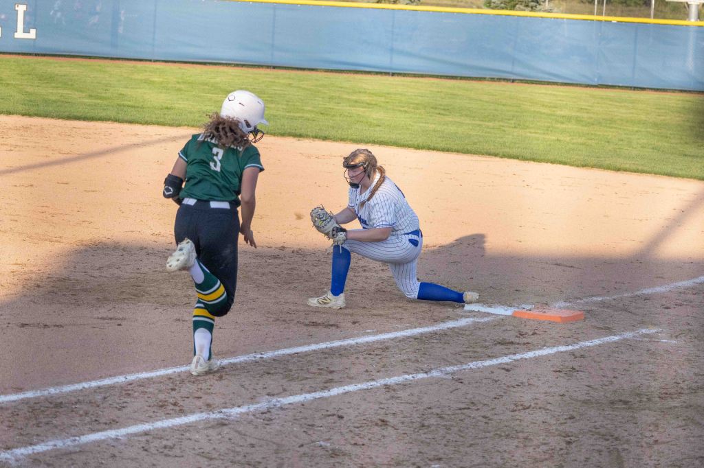 A softball player in blue and white stretches to catch the ball at first base while an opposing player in green runs towards her.