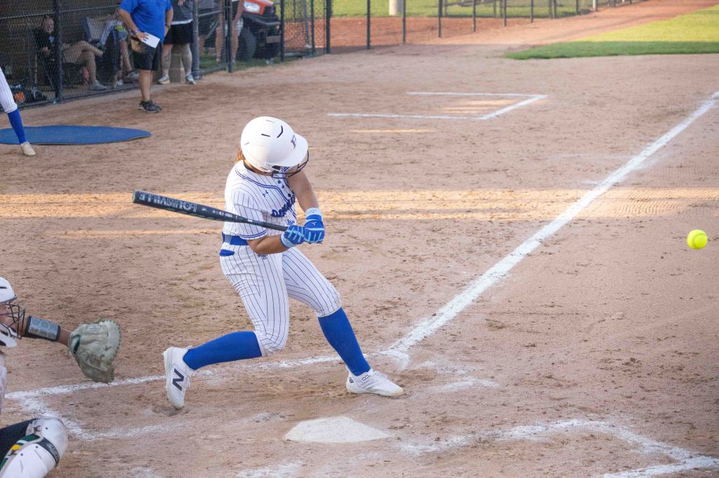A young female softball player in a white and blue uniform swings a bat at a softball during a game, with a catcher in the foreground and spectators in the background.