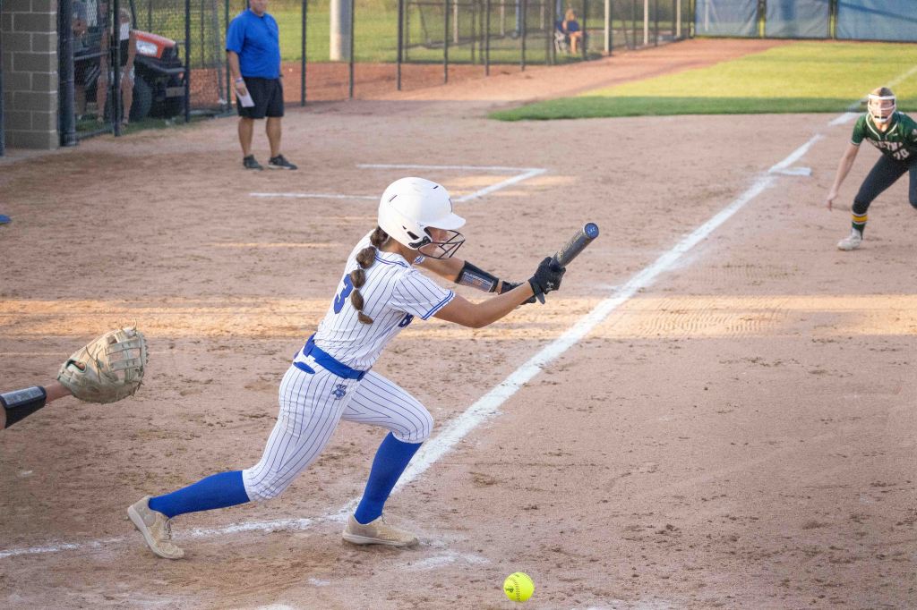 A young female softball player in a white and blue uniform is bunting a ball on a dirt field during a game. In the background, another player is ready in a green uniform, and a coach is observing from the sidelines.
