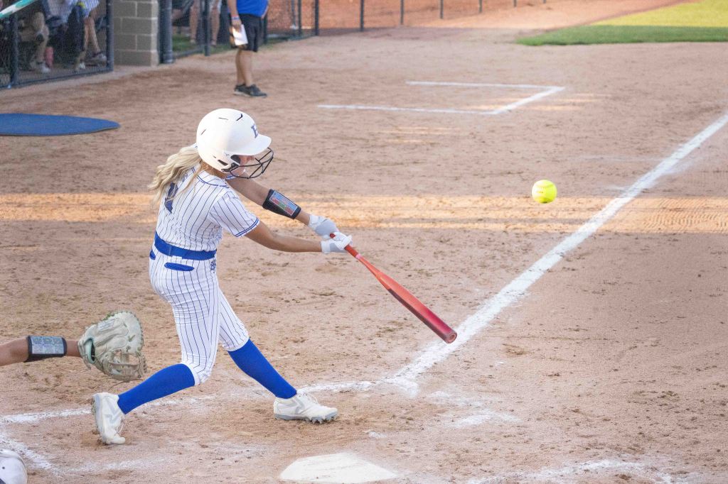 A young female softball player swinging a bat at a pitched ball on a dirt field, with a catcher in the foreground.