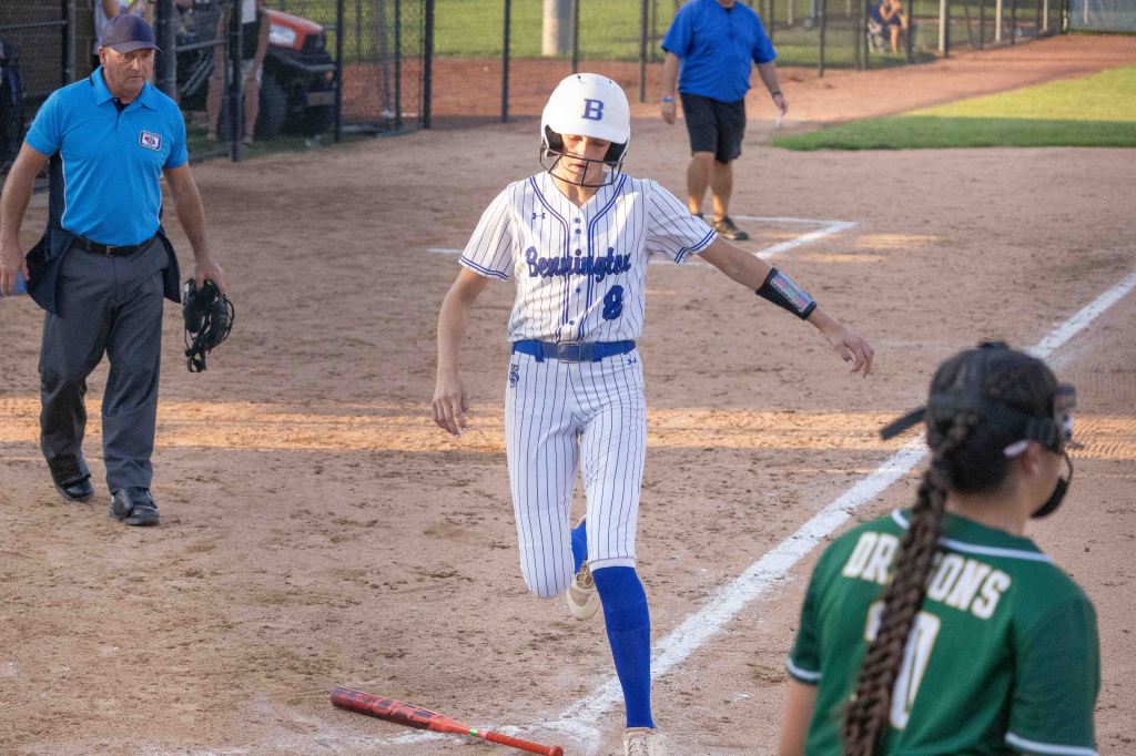 A young softball player in a blue and white striped uniform (#8) crosses home plate during a game, while an umpire and other players are in the background.