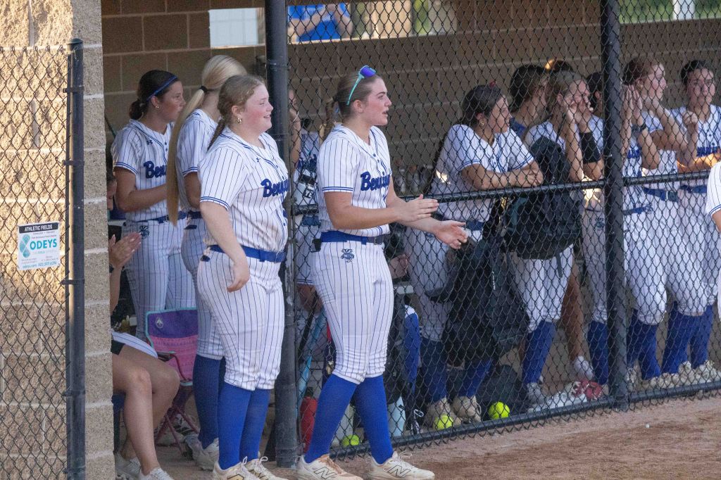 A softball team in a dugout, with players wearing blue and white uniforms, cheering and clapping for their teammates on the field.