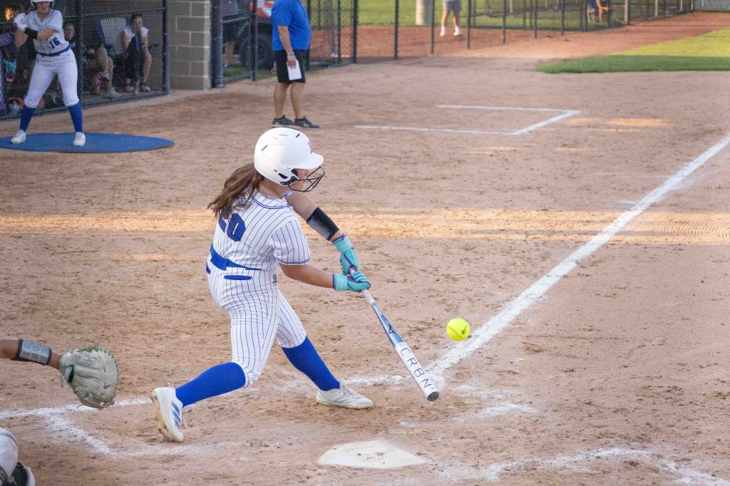 A young female softball player in a white and blue uniform swings her bat at a yellow softball during a game.