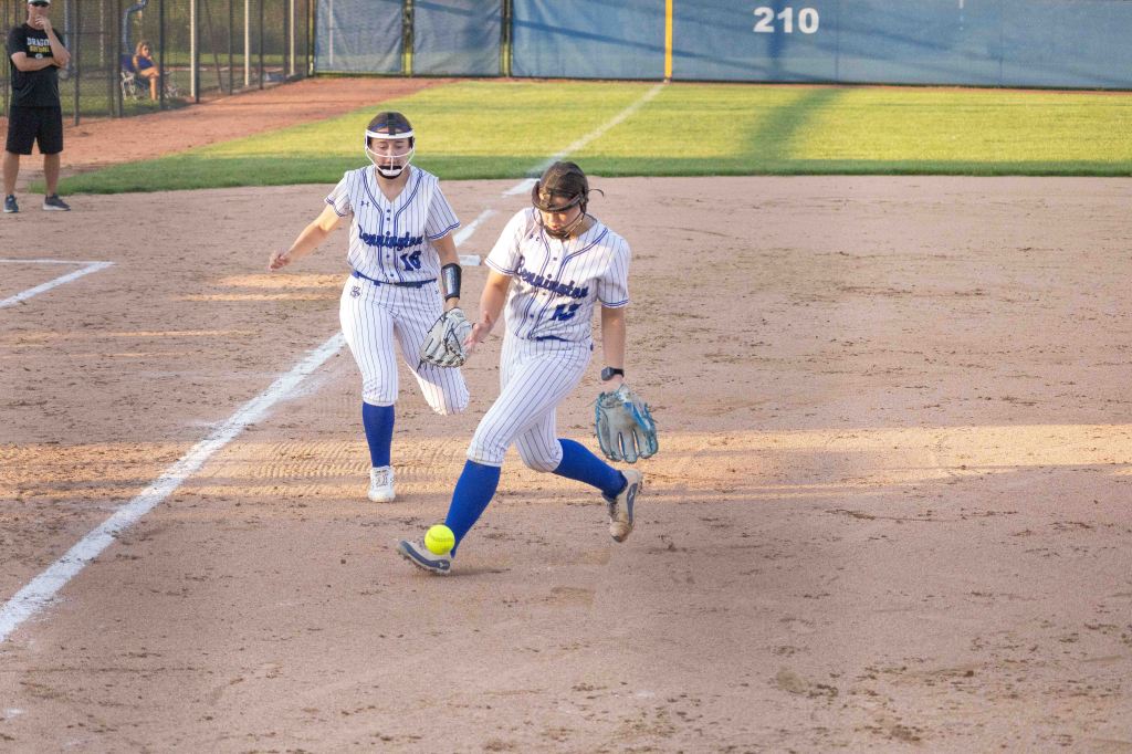 Two female softball players in matching striped uniforms are on the field, with one player reaching for a yellow softball on the ground while the other approaches from behind.
