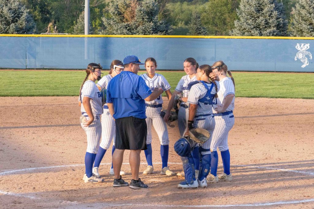 A softball coach discusses strategy with a group of female players on the field during a game.