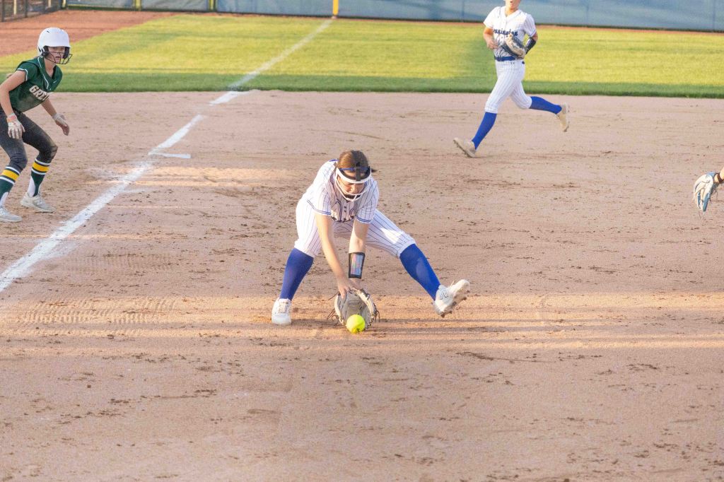 A softball player in a striped jersey and blue pants fields a ground ball on a sandy infield, while another player in a green uniform looks on.