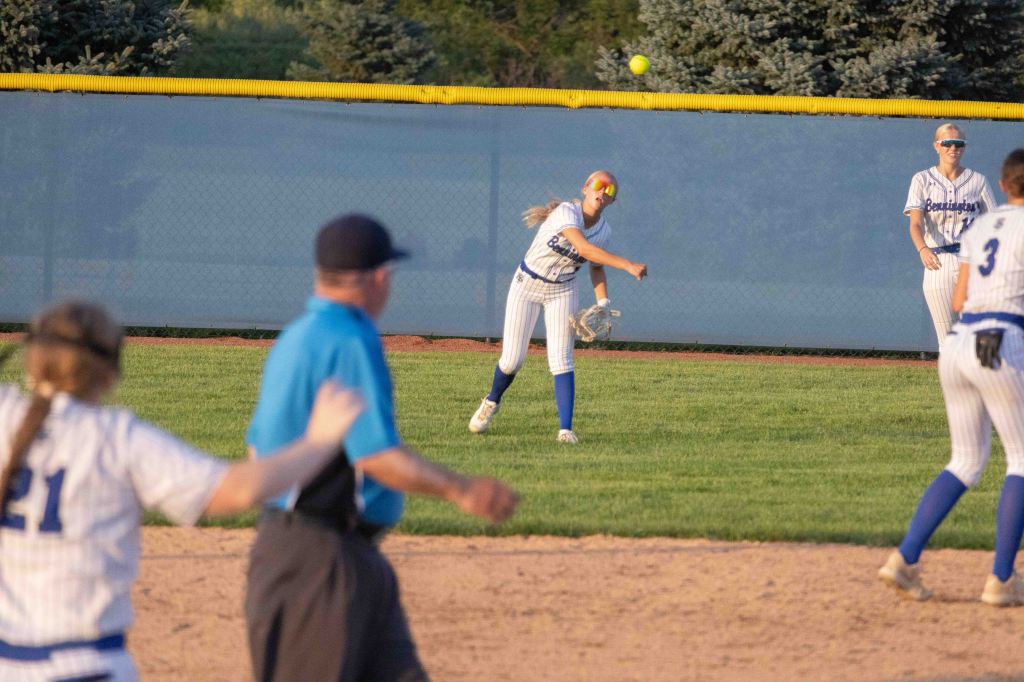 A young female softball player throws a ball while positioned in the outfield, with an umpire in the foreground and other players watching in the background.