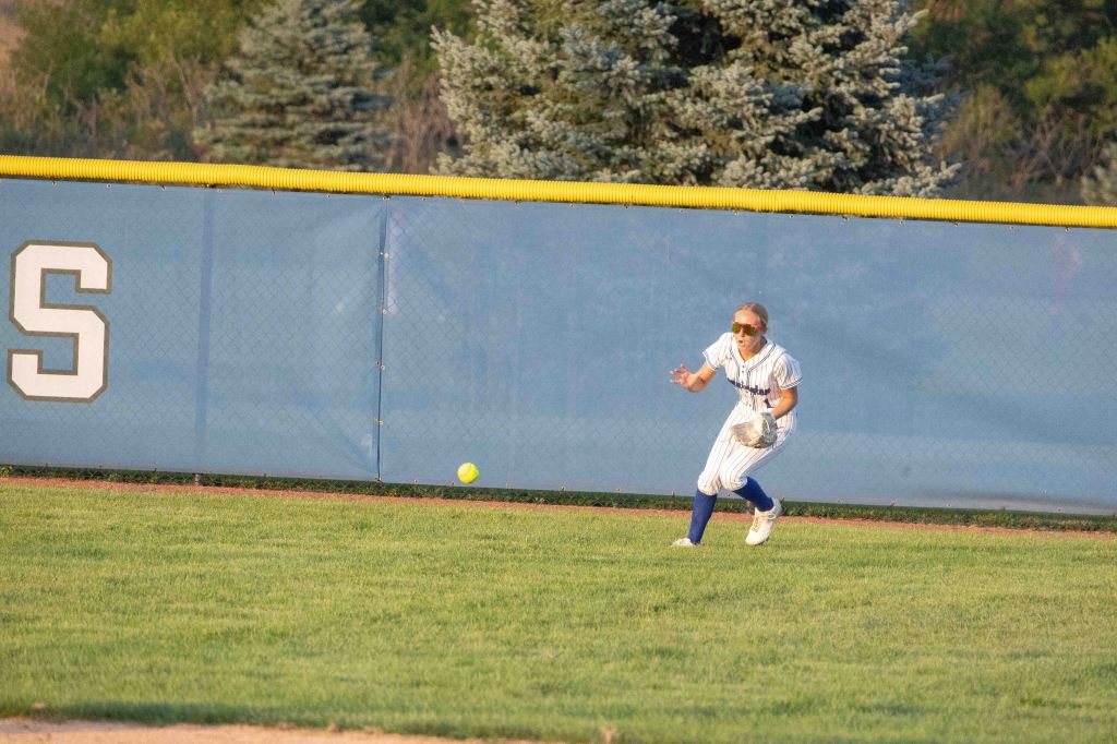 A softball player in a striped uniform and sunglasses is running towards a ball near the outfield fence.