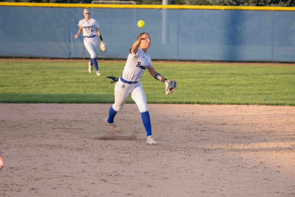 A softball player in a striped uniform throws a ball during a game, while another player is positioned in the background.