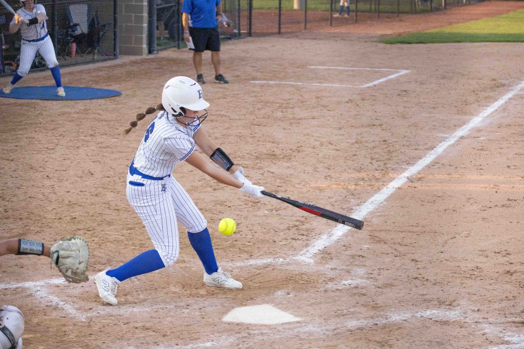 A young female softball player swings her bat at a pitched softball during a game on a dirt field.