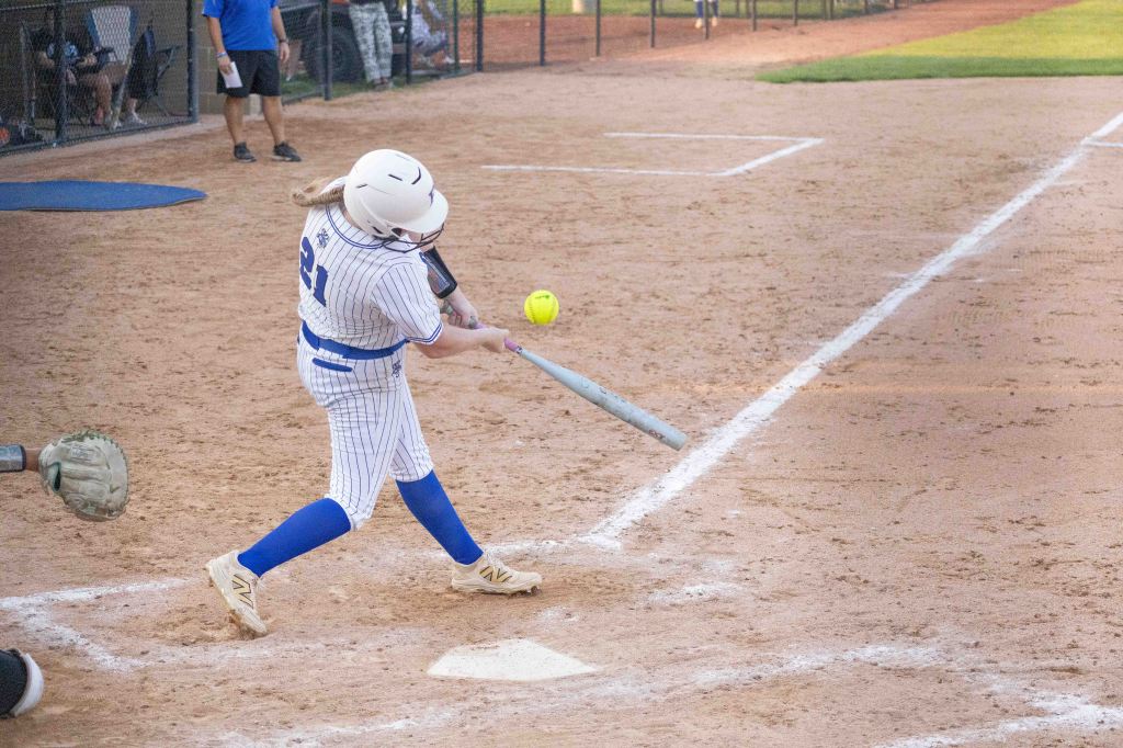 A young female softball player wearing a white and blue striped uniform swings a bat at a yellow softball during a game on a dirt field.