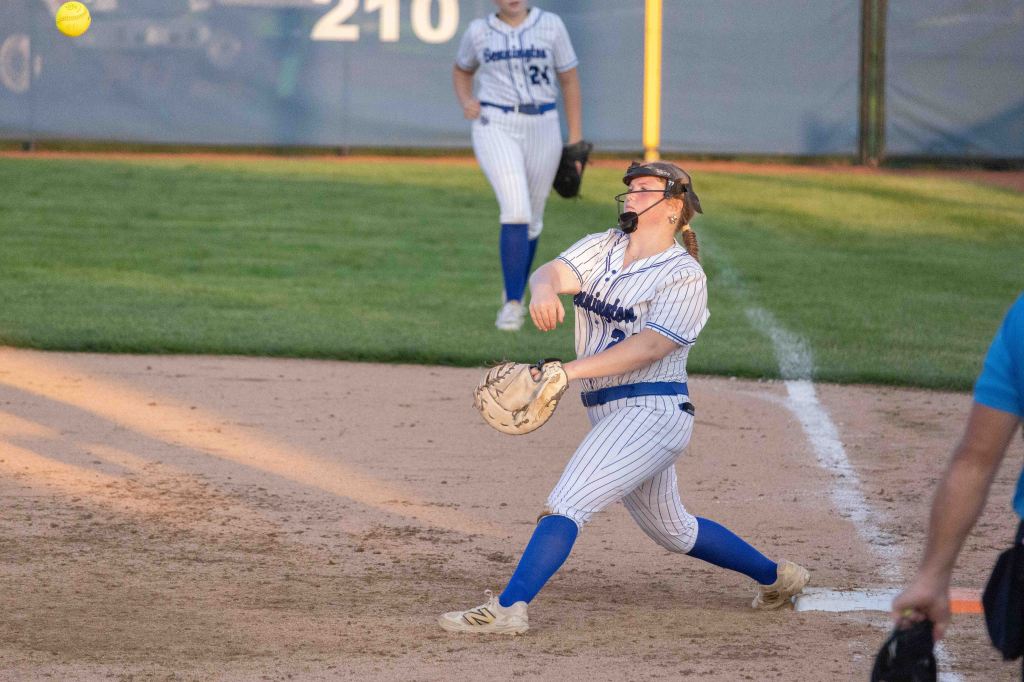 A softball player in a striped uniform prepares to pitch on a dirt field with a yellow softball in the air.