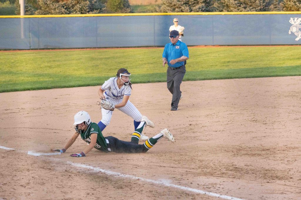 A softball player in a green uniform slides into second base while another player in a white and blue striped uniform prepares to catch the ball. An umpire watches the play unfold in the background.