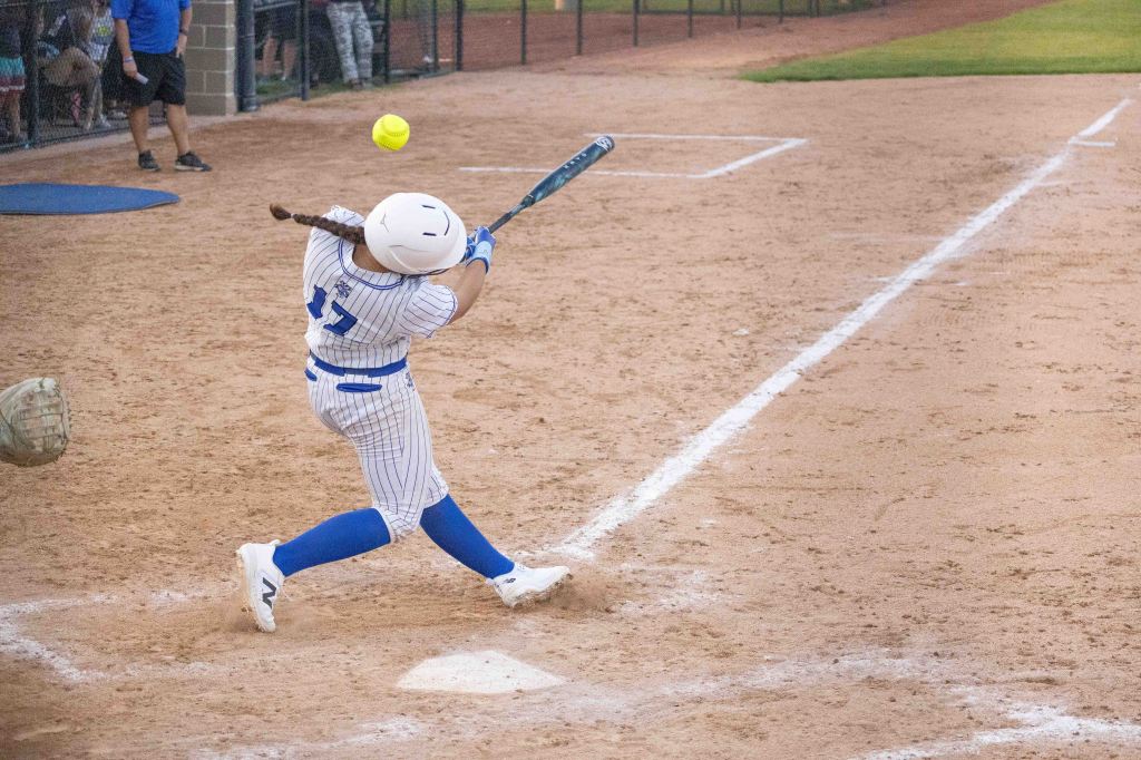 A young female softball player swings a bat to hit a yellow softball during a game on a dirt field.