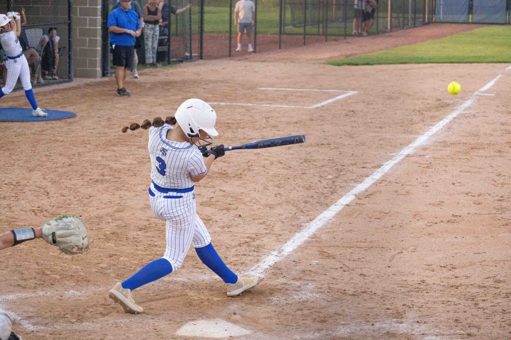 A young female softball player in a striped uniform swings her bat to hit a yellow softball during a game, with spectators in the background.