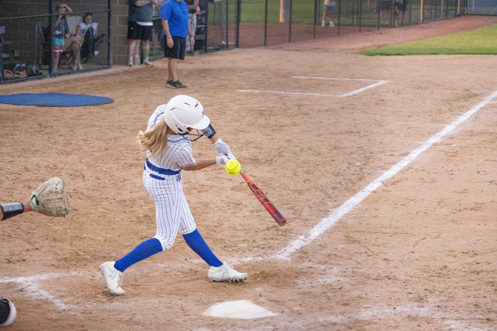 A young girl in a baseball uniform swings a bat at a softball on a dirt field, with spectators in the background.