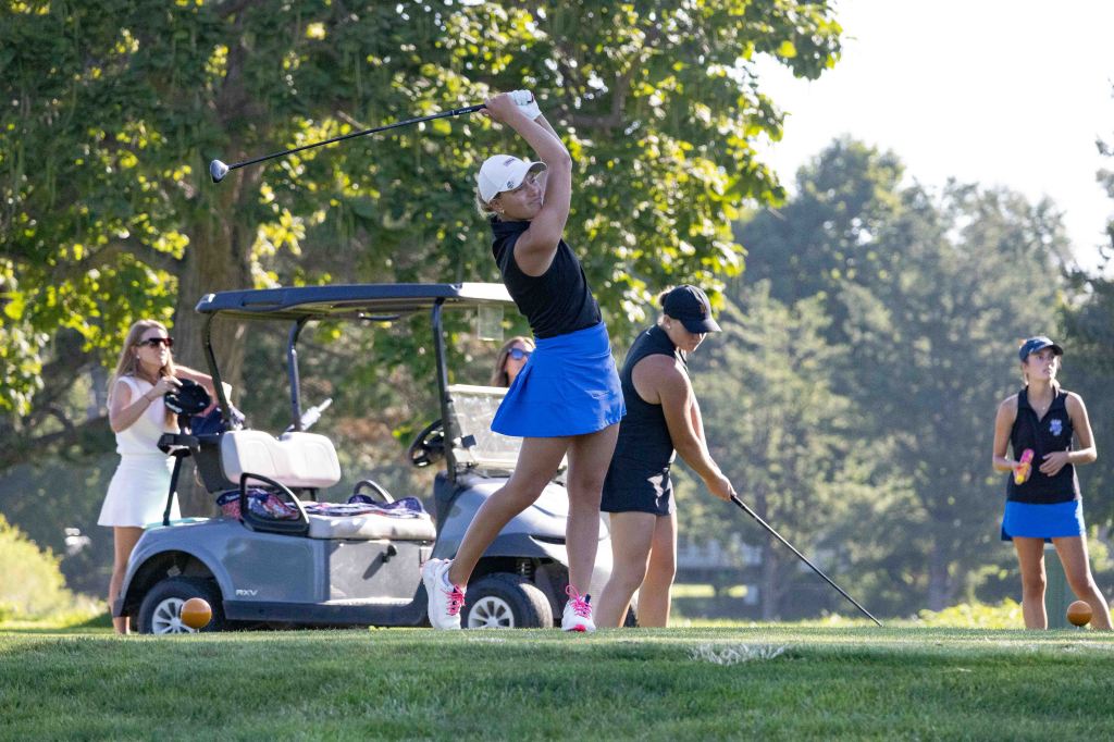 Four women playing golf on a sunny day, with one swinging a club and others observing, near a golf cart.