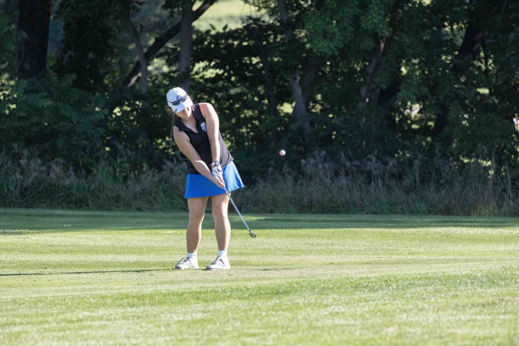 A female golfer in a black sleeveless top and blue skirt swings a club on a sunny golf course, with trees in the background.