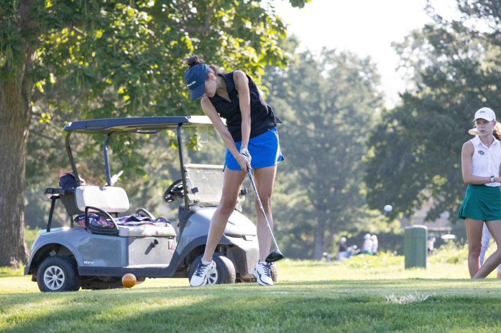 A female golfer in a black sleeveless top and blue shorts, preparing to swing her club on a grassy golf course, with a golf cart in the background and another female golfer watching.