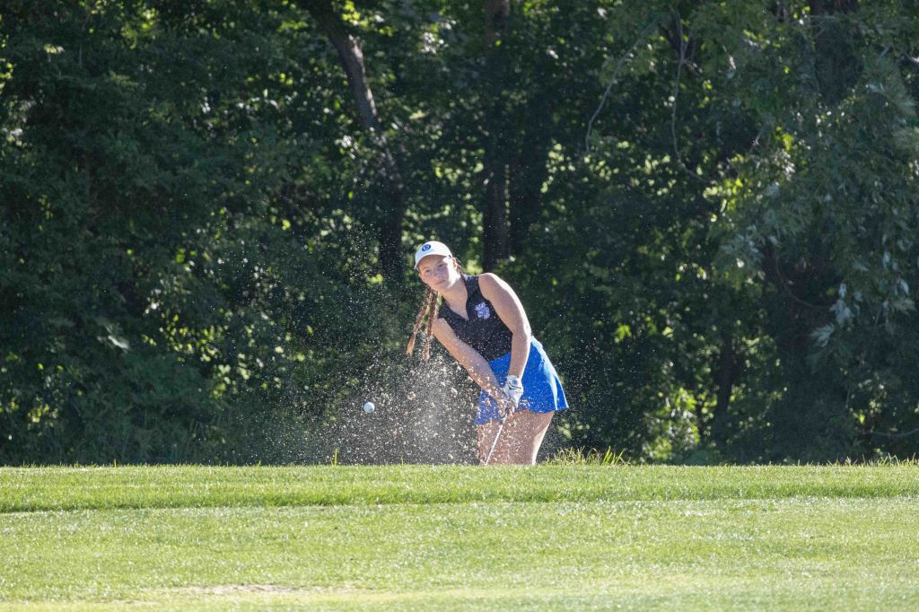A female golfer hitting a sand shot from a bunker on a golf course, with sand flying around her as she swings.