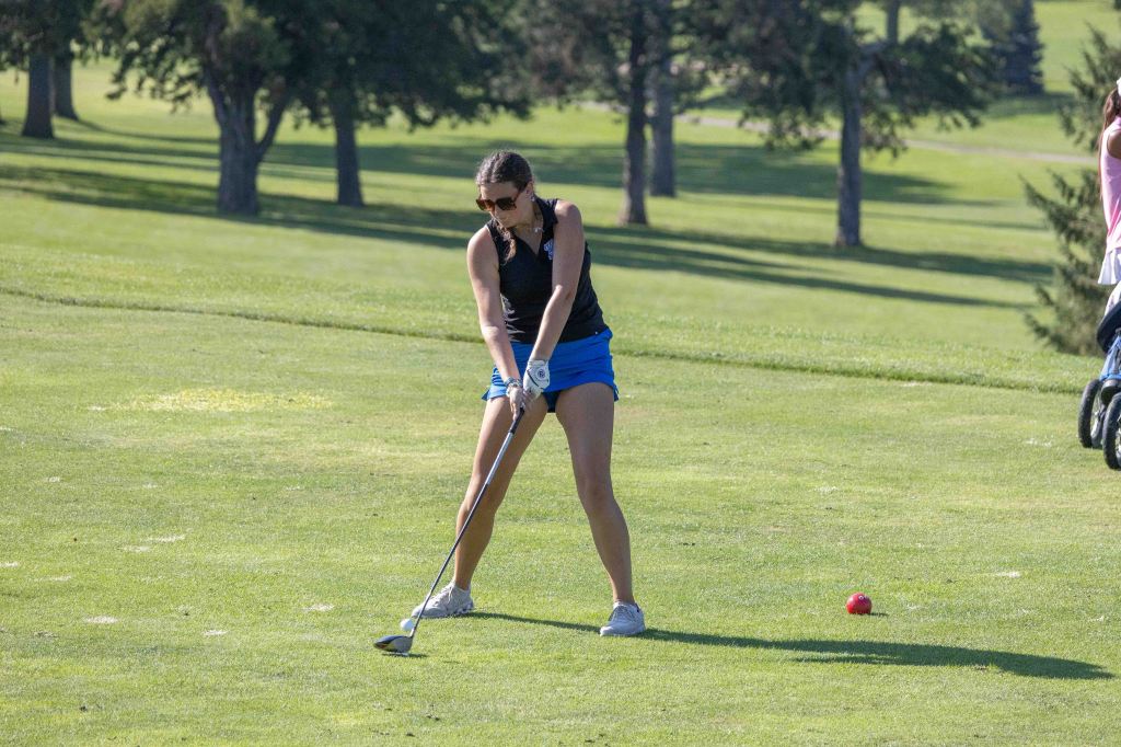 A woman preparing to swing a golf club on a sunny day at a golf course, wearing a black sports top and blue shorts.