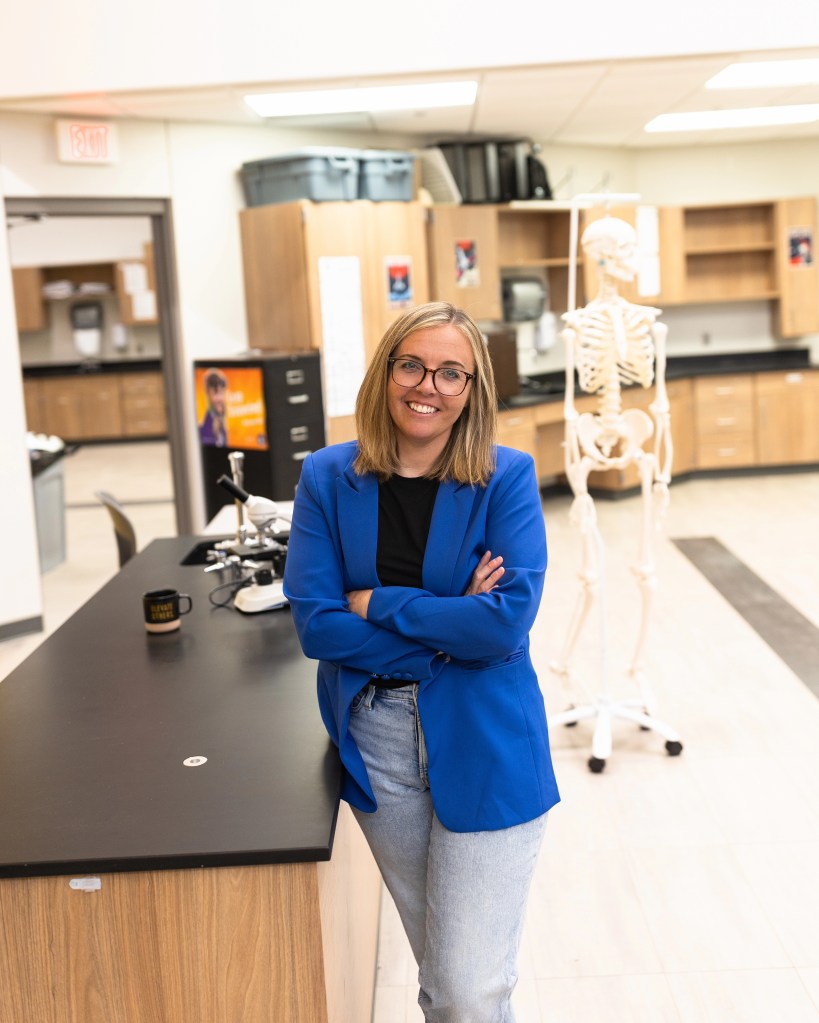 A smiling woman wearing a blue blazer and glasses stands with her arms crossed at a science lab workstation, featuring a skeleton model in the background.