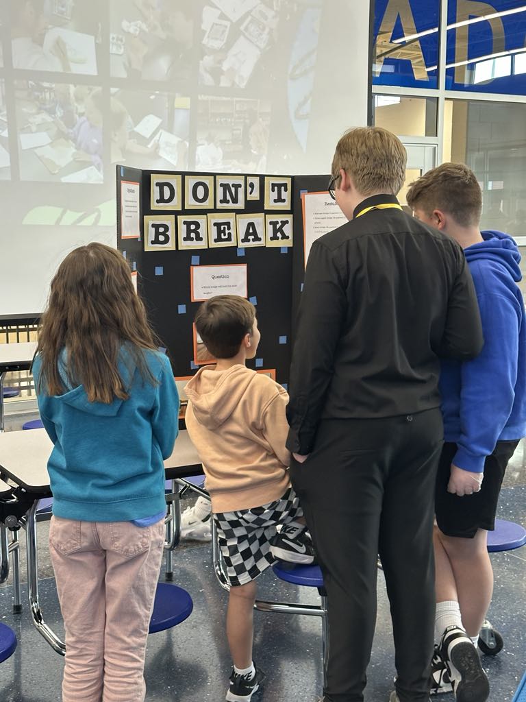 A group of four children and a teenager gathered around a science project display board that reads 'DON'T BREAK.' The display features information on an experiment, with background visuals of students working on similar projects.