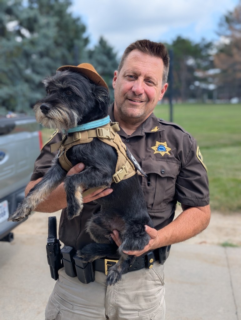 A police officer in uniform holding a small dog wearing a harness and a hat, standing outdoors with trees in the background.