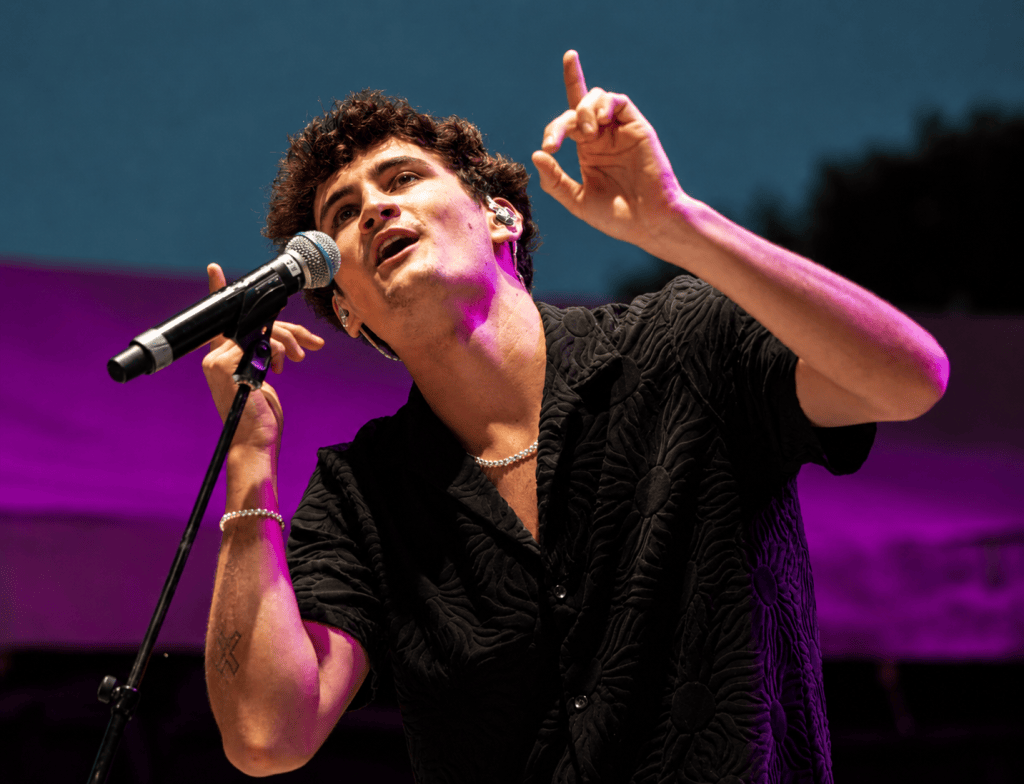 A male singer passionately performing on stage with a microphone, dressed in a black patterned shirt, gesturing expressively.