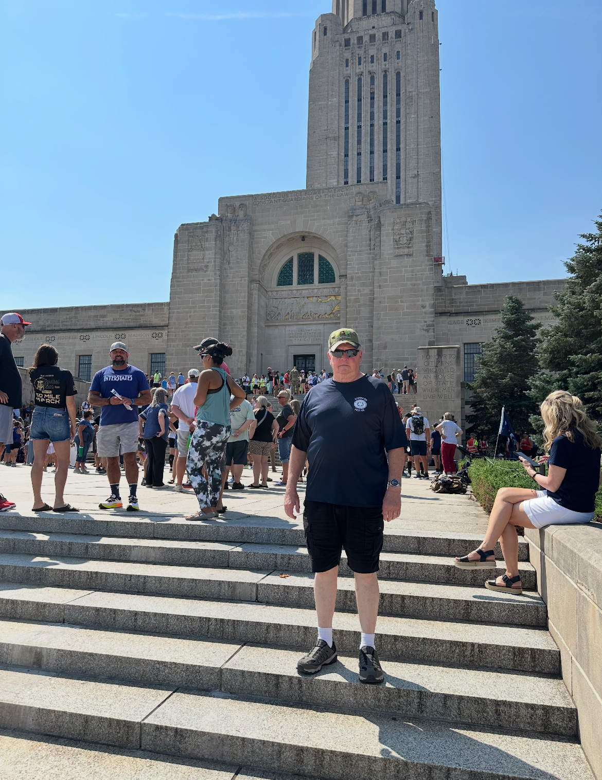 A man standing on concrete steps in front of a large historic building, with crowds of people gathered around in a sunny outdoor setting.