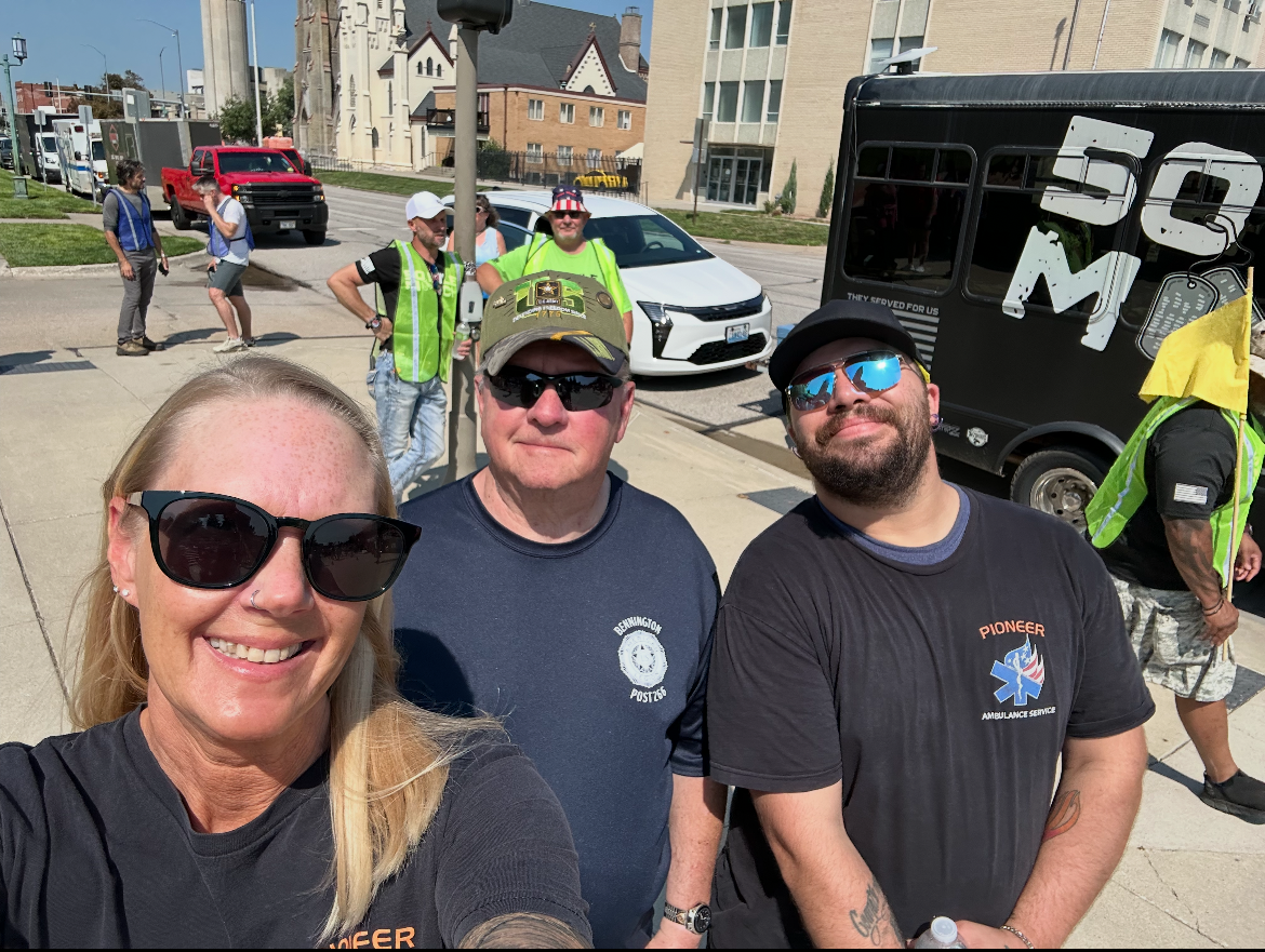 Three people posing for a selfie outdoors on a sunny day, with a line of vehicles in the background. The person on the left has long blonde hair and sunglasses, the middle person is wearing a dark shirt and sunglasses, and the person on the right has a beard and is also wearing sunglasses. Other individuals can be seen in the background chatting and standing near vehicles.