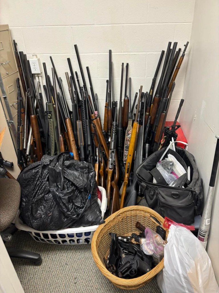 A cluttered storage room filled with numerous firearms leaning against the wall, including rifles and handguns, alongside baskets and bags containing additional items.