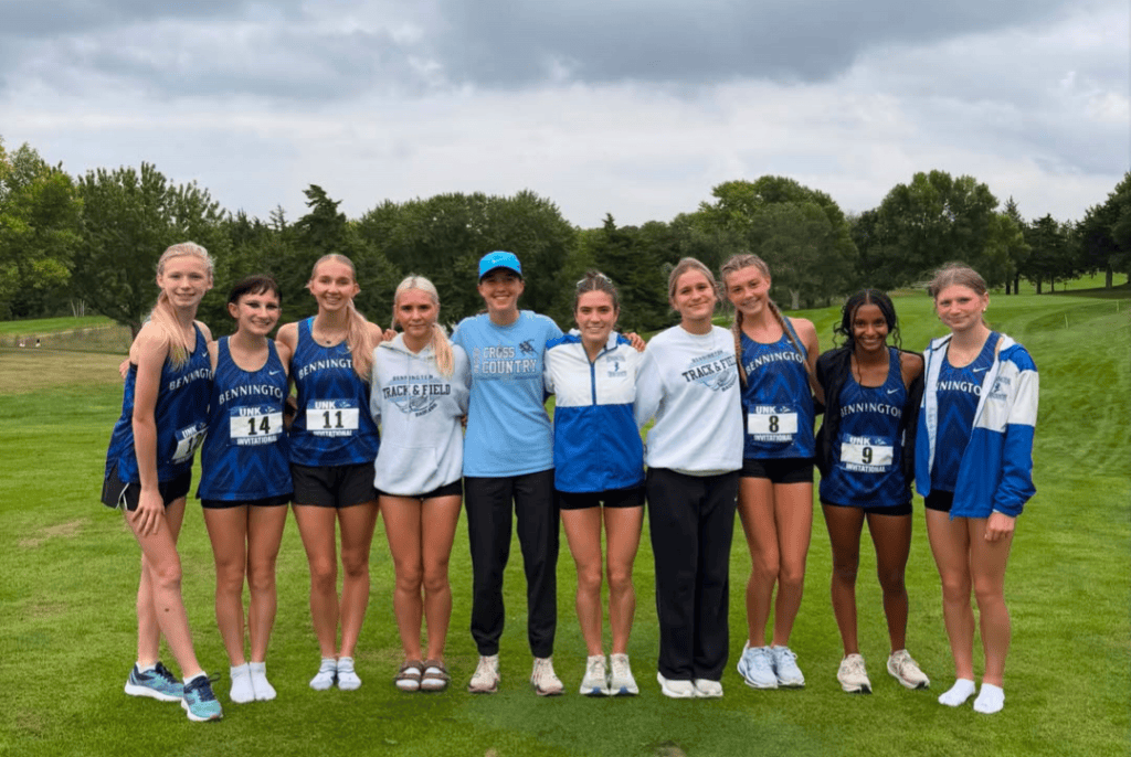 Group photo of a girls' cross country team posing on a grassy field, wearing matching blue and black athletic uniforms.