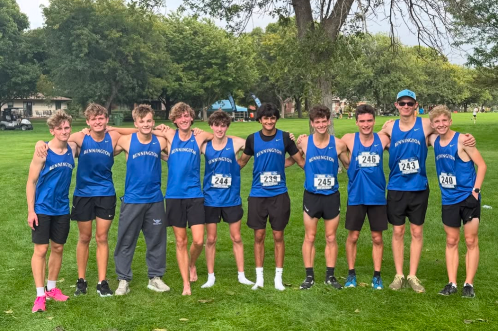 A group of ten male cross-country runners in blue uniforms stands together on a grassy field, smiling for the camera.