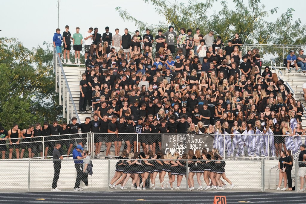 A large crowd of students dressed in black shirts gathered in a stadium, some holding a banner that reads 'it's DARK out, hope you can SEE.' Cheerleaders are at the front, and the stands are filled with supporters.