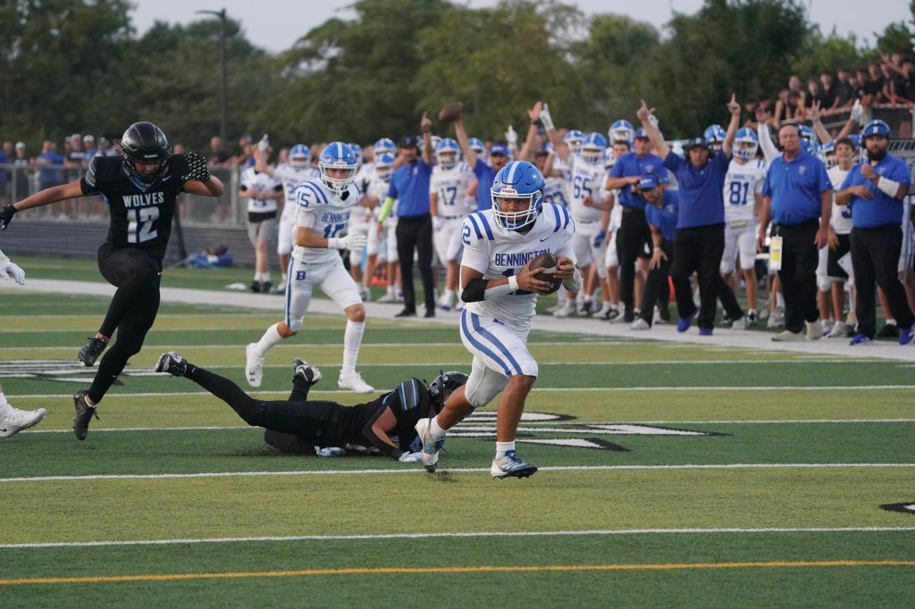 A football player in a blue and white uniform runs with the ball while avoiding a tackle from a player in a black uniform on the field. In the background, spectators cheer for the team.