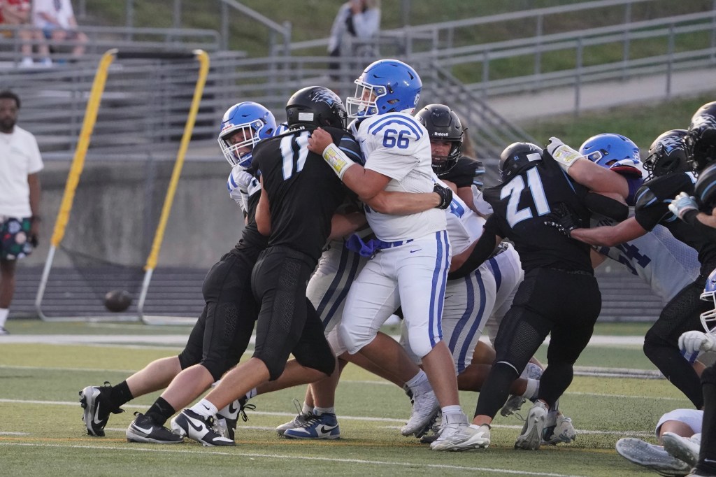 Football players engaged in a tackle during a game, with one player in a blue jersey and another in a black jersey being pushed together while other players are in action in the background.