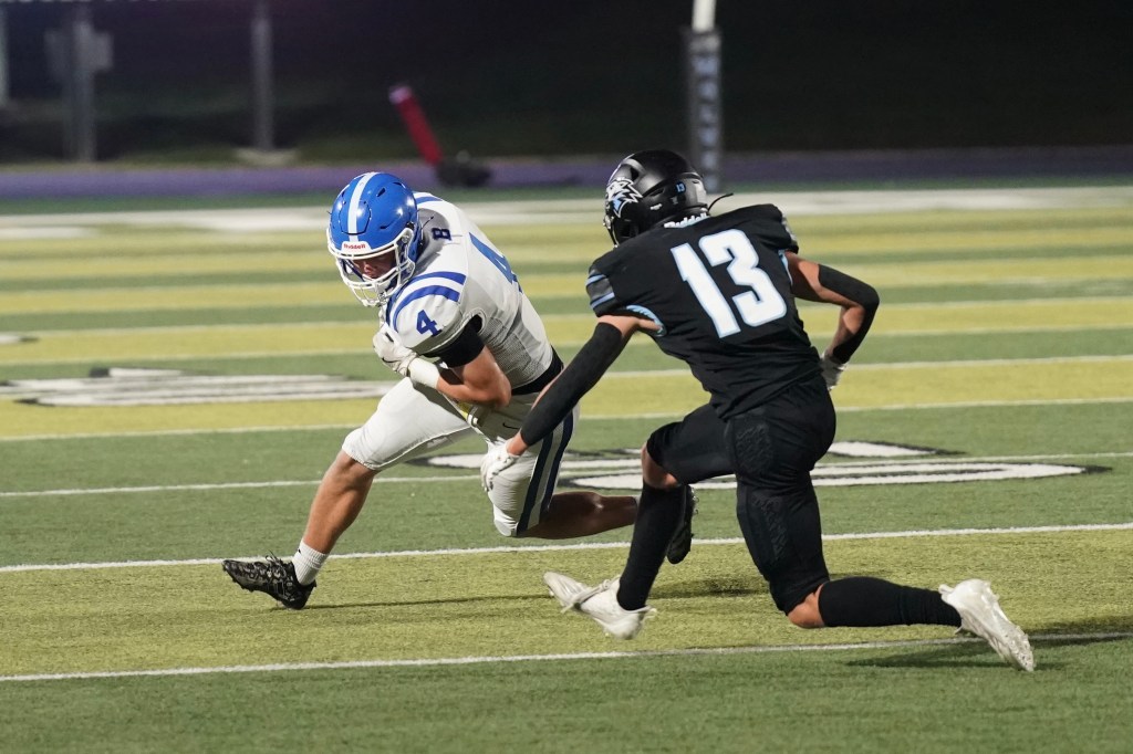 A football player in a white uniform with blue accents, number 4, evades a defender in a black uniform with number 13 on a football field under stadium lights.