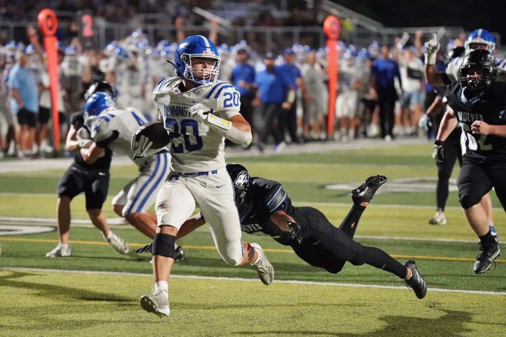 A high school football player in a white jersey with blue stripes, wearing the number 20, runs with the ball while a defender in black attempts to tackle him. The background shows other players in motion and spectators watching the game.