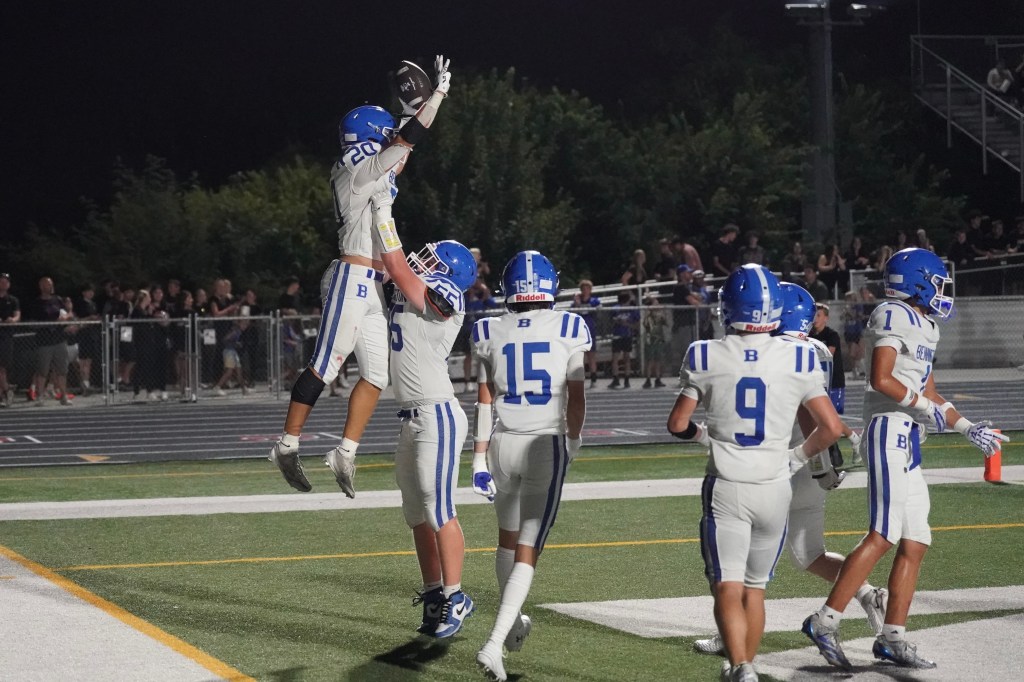 A youth football team celebrating a touchdown with one player being lifted in the air by a teammate, surrounded by other teammates on the field at night.