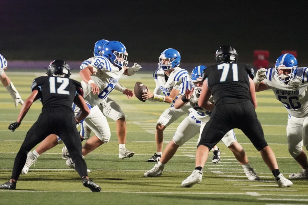 High school football players in action during a night game, with a ball carrier surrounded by teammates in blue uniforms and opponents in black.